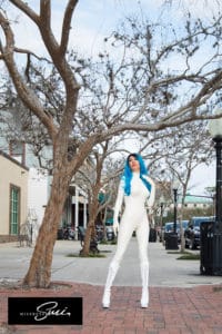Woman with blue hair in a white bodysuit standing outdoors near a tree.