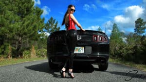 Woman in red top and black leather pants standing by a black car outdoors.
