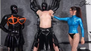 A woman in a blue latex outfit interacts with two masked individuals in a BDSM scene.