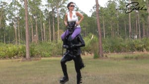 Woman in fetish outfit standing on a man in an outdoor forest setting.